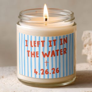 May include: A lit candle in a clear glass jar. The candle has a cream-colored wax and a label with the text "I LEFT IT IN THE WATER" in red, with blue and white stripes. The date "4.26.26" is below the text.
