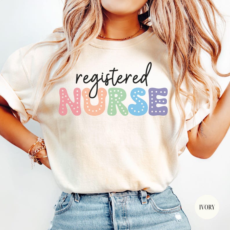 May include: A white t-shirt with the text "registered NURSE" in colorful letters with white polka dots. The letters are in a rainbow color scheme. The shirt is being worn by a person with long blonde hair and is paired with blue jeans.
