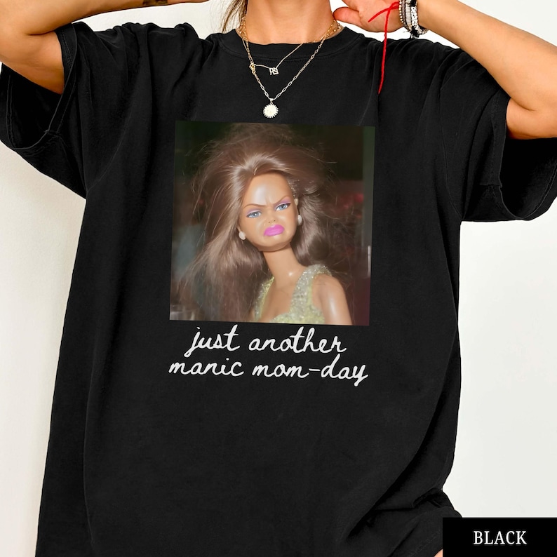 May include: Black t-shirt featuring a meme of a doll with a displeased expression. The doll is in a square image with the text "just another manic mom-day" in a handwritten font. The shirt is a basic crew neck.