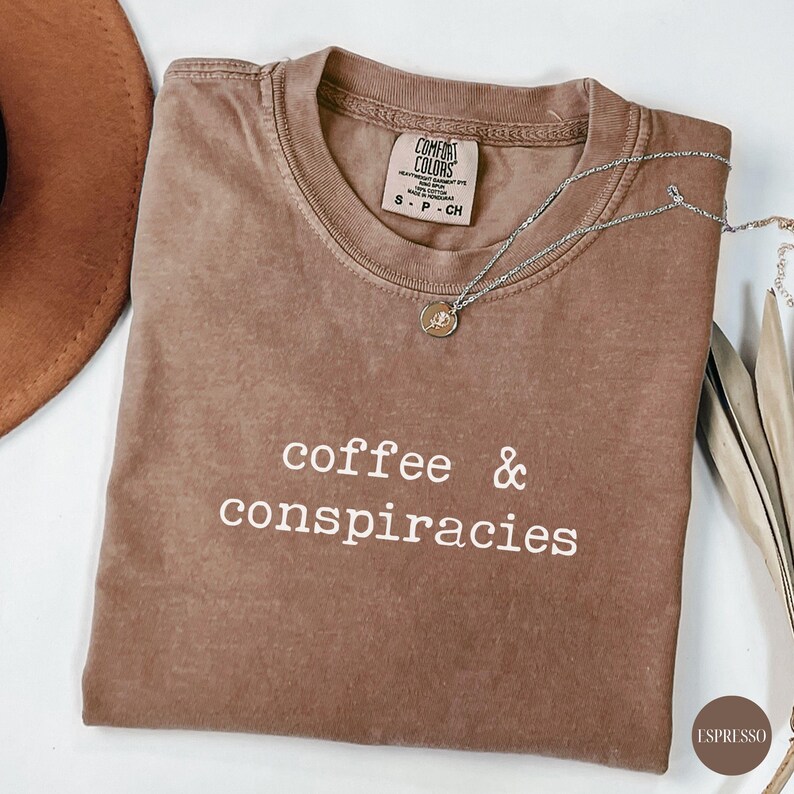 K&ouml;nnte beinhalten: Espressofarbenes T-Shirt mit den wei&szlig;en Worten "coffee & conspiracies". Eine silberne Halskette mit einem kleinen Anh&auml;nger liegt auf dem Shirt. Das Shirt ist gefaltet und hat ein Etikett am Ausschnitt.