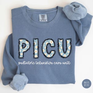 Puede incluir: Una sudadera azul con un estampado de margaritas blancas que deletrea "PICU" y el texto "unidad de cuidados intensivos pediátricos".