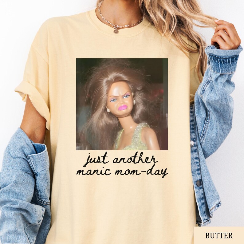 May include: Butter-colored t-shirt featuring a framed image of a doll with a displeased expression. The text "just another manic mom-day" is printed below the image in a handwritten style. The shirt has rolled-up sleeves.