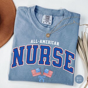 Op de afbeelding: Blauw spijkerbroekkleurig T-shirt met de woorden "ALL-AMERICAN NURSE" in blauwe en rode letters. Twee Amerikaanse vlaggen zijn onder de tekst gedrukt. Een gouden ketting met een hanger is over het shirt gedrapeerd.
