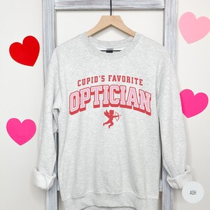 Puede incluir: Una sudadera gris con el texto "Cupid's Favorite Optician" en letras rosa y rojas. Un peque&ntilde;o cupido rojo con una flecha est&aacute; debajo del texto.