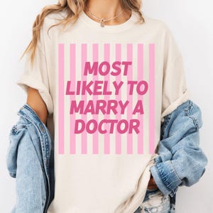 Op de afbeelding: Cr&egrave;mekleurig T-shirt met roze verticale strepen en de roze tekst "MOST LIKELY TO MARRY A DOCTOR". Het shirt heeft opgerolde mouwen en wordt gedragen met een spijkerjasje over de schouders.