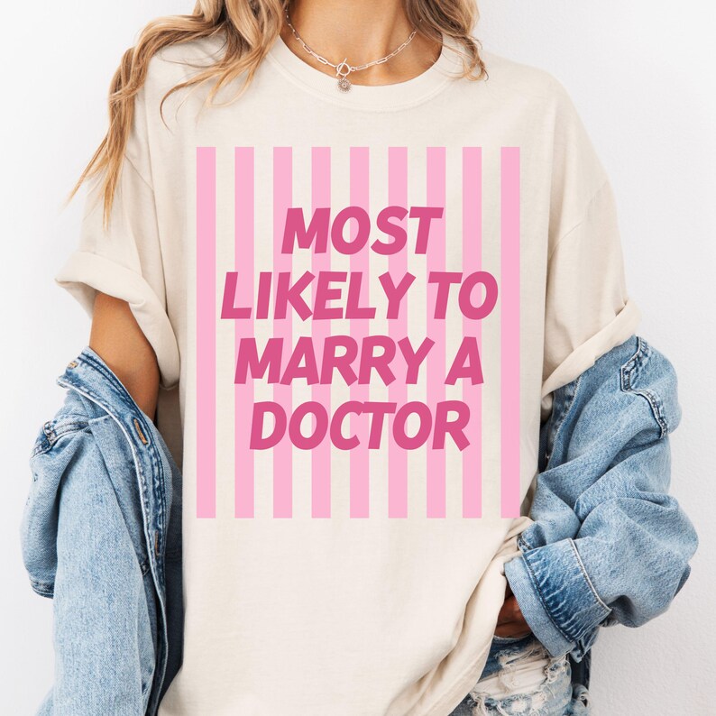 Op de afbeelding: Cr&egrave;mekleurig T-shirt met roze verticale strepen en de roze tekst "MOST LIKELY TO MARRY A DOCTOR". Het shirt heeft opgerolde mouwen en wordt gedragen met een spijkerjasje over de schouders.