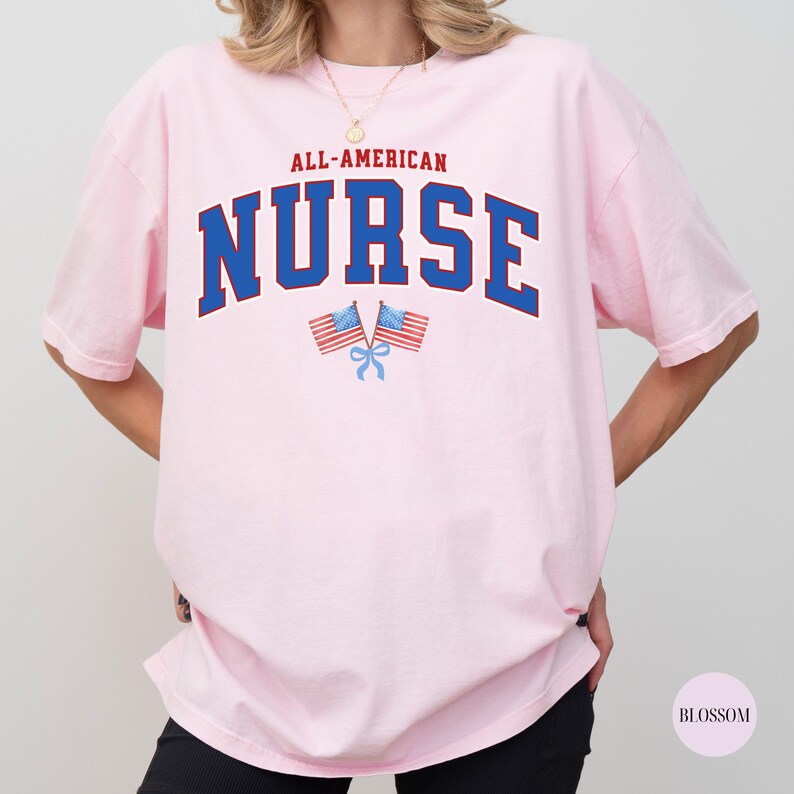 Op de afbeelding: Lichtroze T-shirt met de tekst "ALL-AMERICAN NURSE" in blauw en rood. Twee kleine Amerikaanse vlaggen staan eronder. Het shirt heeft een casual stijl.