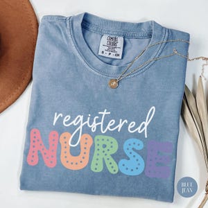 May include: A light blue t-shirt with the text "registered NURSE" in white with the letters in rainbow colors and white polka dots. The shirt is made of a soft, comfortable material.