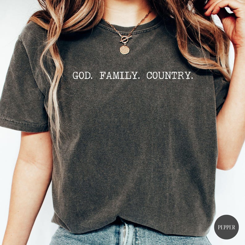 May include: Dark gray t-shirt with the text "GOD. FAMILY. COUNTRY." in white block letters. The shirt has short sleeves and a slightly faded appearance. The model is wearing a gold necklace.