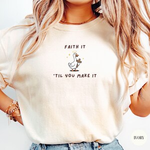 May include: Ivory-colored t-shirt featuring a cartoon goose holding a cross with a halo above its head. The text "FAITH IT" is above the goose, and "'TIL YOU MAKE IT" is below. The shirt is a comfortable, casual style.