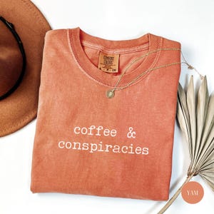 K&ouml;nnte beinhalten: Ein korallenfarbenes Comfort Colors T-Shirt mit den wei&szlig;en Worten "coffee & conspiracies". Eine goldene Halskette mit Anh&auml;nger liegt auf dem Shirt. Ein brauner Hut und ein getrocknetes Palmblatt sind ebenfalls im Bild.