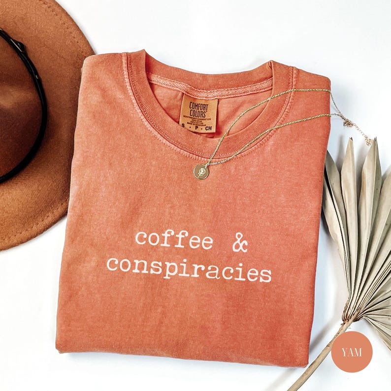 K&ouml;nnte beinhalten: Ein korallenfarbenes Comfort Colors T-Shirt mit den wei&szlig;en Worten "coffee & conspiracies". Eine goldene Halskette mit Anh&auml;nger liegt auf dem Shirt. Ein brauner Hut und ein getrocknetes Palmblatt sind ebenfalls im Bild.