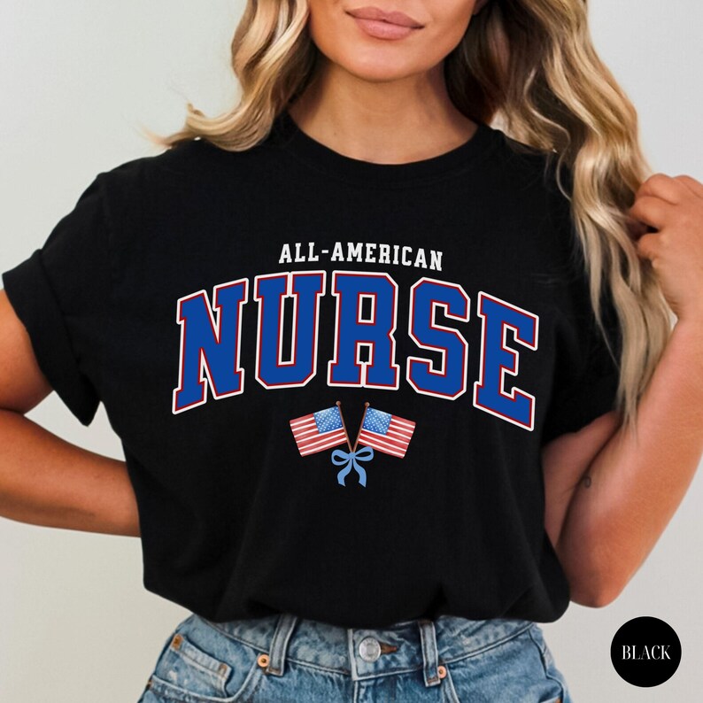 Op de afbeelding: Zwart T-shirt met de tekst "ALL-AMERICAN NURSE" in wit en blauw, met twee Amerikaanse vlaggen en een blauw lint. Het shirt heeft een ronde hals.