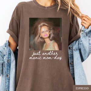 May include: Espresso-colored t-shirt featuring a square image of a doll with a displeased expression. The text "just another manic mom-day" is printed below the image in a handwritten style. The shirt has a relaxed fit.