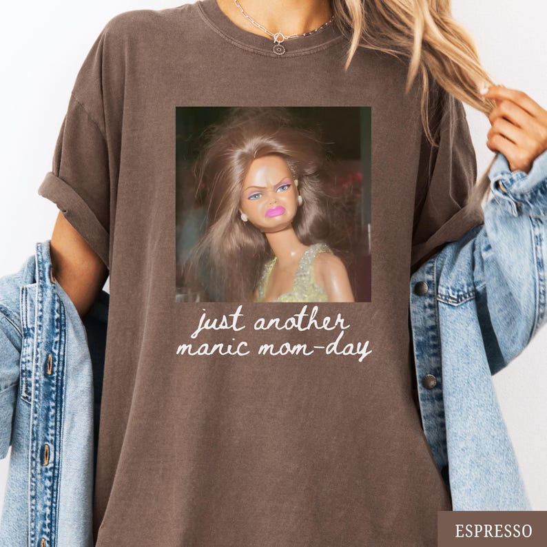 May include: Espresso-colored t-shirt featuring a square image of a doll with a displeased expression. The text "just another manic mom-day" is printed below the image in a handwritten style. The shirt has a relaxed fit.