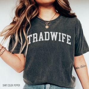 Puede incluir: Una camiseta gris oscuro con la palabra "TRADWIFE" impresa en blanco en la parte delantera. La camiseta est&aacute; hecha de un material suave y c&oacute;modo y tiene un corte holgado.