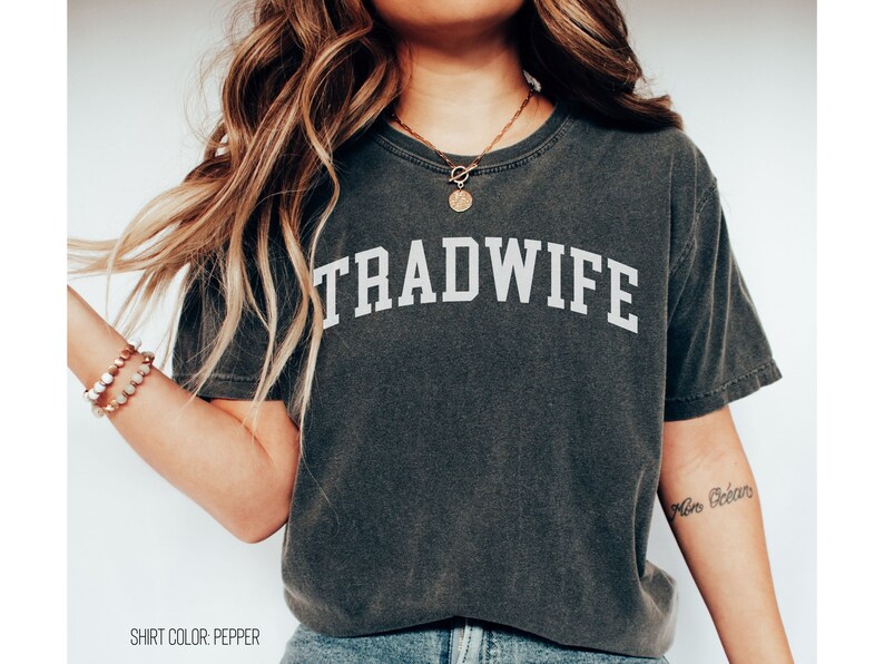 Puede incluir: Una camiseta gris oscuro con la palabra "TRADWIFE" impresa en blanco en la parte delantera. La camiseta est&aacute; hecha de un material suave y c&oacute;modo y tiene un corte holgado.