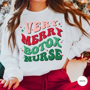 Puede incluir: Sudadera blanca con cuello redondo con el texto "VERY MERRY BOTOX NURSE" en rosa, rojo y verde, con detalles de estrellas. La sudadera se combina con pantalones rojos.