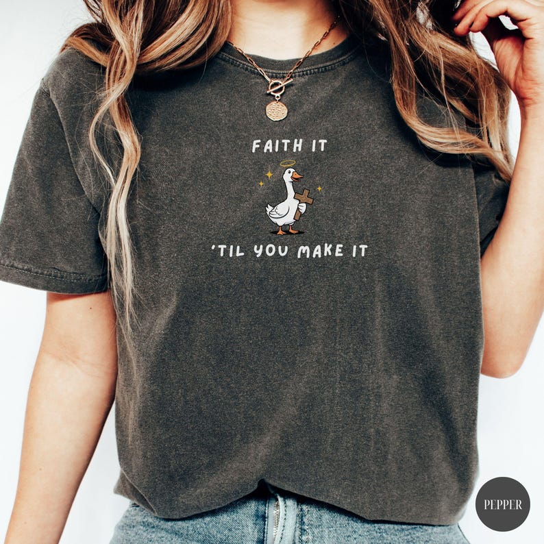 May include: Dark gray t-shirt with the text "FAITH IT 'TIL YOU MAKE IT". The shirt features a cartoon goose with a halo holding a cross. The shirt is a comfortable, casual style.