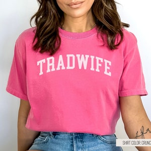 Puede incluir: Una camiseta rosa con el texto "TRADWIFE" en letras blancas. La camiseta es de corte holgado y tiene cuello redondo. El color de la camiseta es "Crunchberry".
