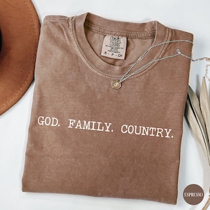 May include: A brown Comfort Colors t-shirt with the words "GOD. FAMILY. COUNTRY." in white. A silver necklace with a pendant rests on the shirt. The shirt is a small size.