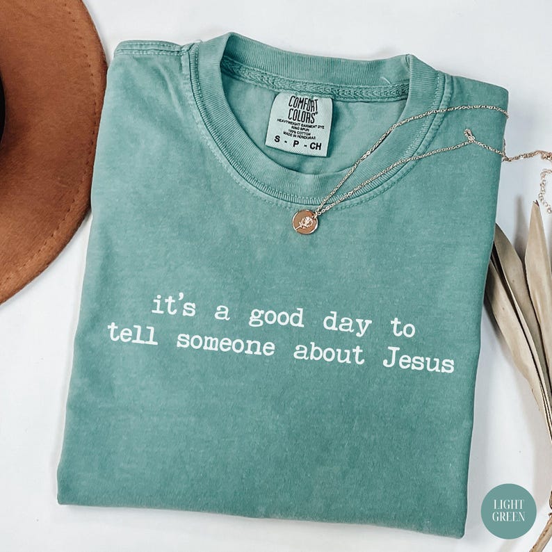 K&ouml;nnte beinhalten: Hellgr&uuml;nes Comfort Colors T-Shirt mit dem wei&szlig;en Text "it's a good day to tell someone about Jesus". Eine goldene Halskette mit Anh&auml;nger liegt auf dem Shirt. Ein brauner Hut ist im Hintergrund teilweise sichtbar.