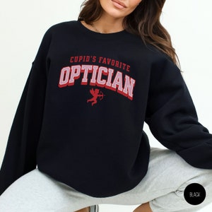 Puede incluir: Sudadera negra con el texto "Cupid's Favorite Optician" en letras rojas y rosas. Un peque&ntilde;o cupido rojo con una flecha est&aacute; por encima del texto.