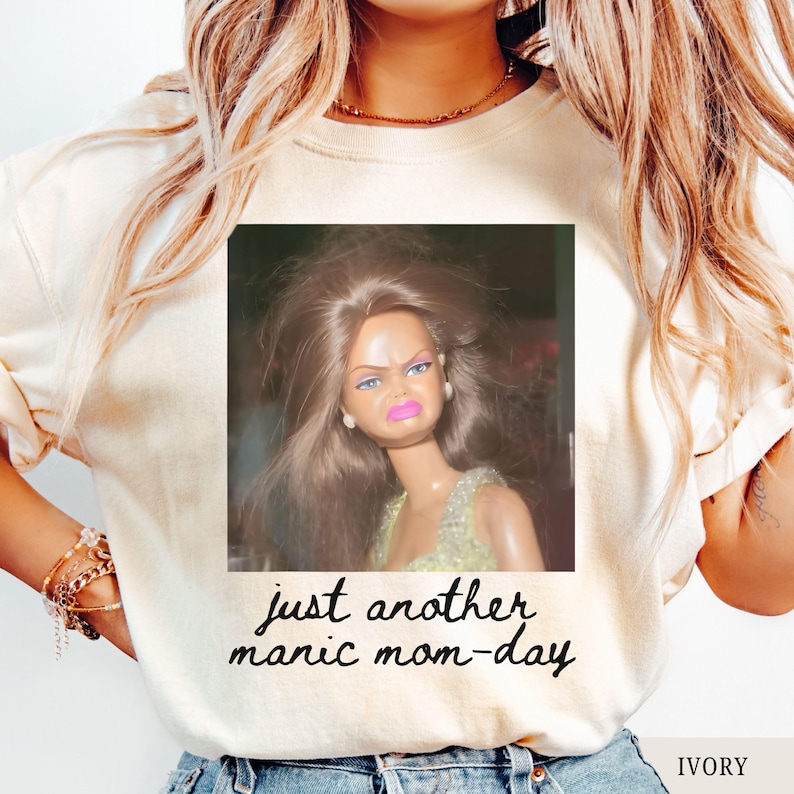 May include: Ivory-colored sweatshirt featuring a square image of a doll with a displeased expression. The text "just another manic mom-day" is printed below the image in a handwritten font. The word "IVORY" is printed at the bottom right.