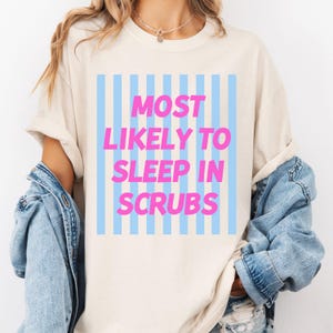 Op de afbeelding: Cr&egrave;mekleurig T-shirt met de tekst "MOST LIKELY TO SLEEP IN SCRUBS" in roze letters. De tekst staat tegen een lichtblauwe en witte gestreepte achtergrond. Onderdeel van een casual kledingcollectie.