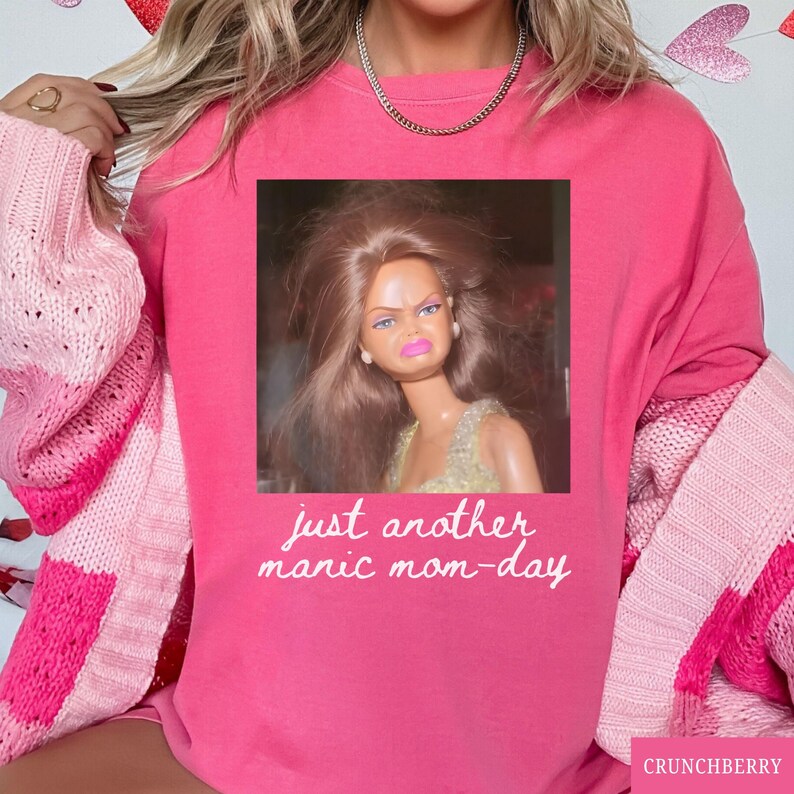 May include: A bright pink sweatshirt with the text "just another manic mom-day" in white script. The sweatshirt features a square image of a doll with a displeased expression. A pink and white sweater is draped over the shoulders.