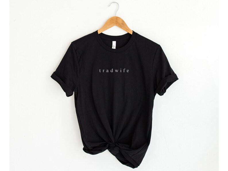 Op de afbeelding: Zwart T-shirt met de tekst "trad wife" in wit gedrukt op de voorkant.
