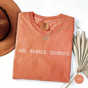 May include: A coral-colored Comfort Colors t-shirt with the words "GOD. FAMILY. COUNTRY." in white. A gold necklace with a pendant rests on the shirt. A brown hat and dried palm leaves are in the background.