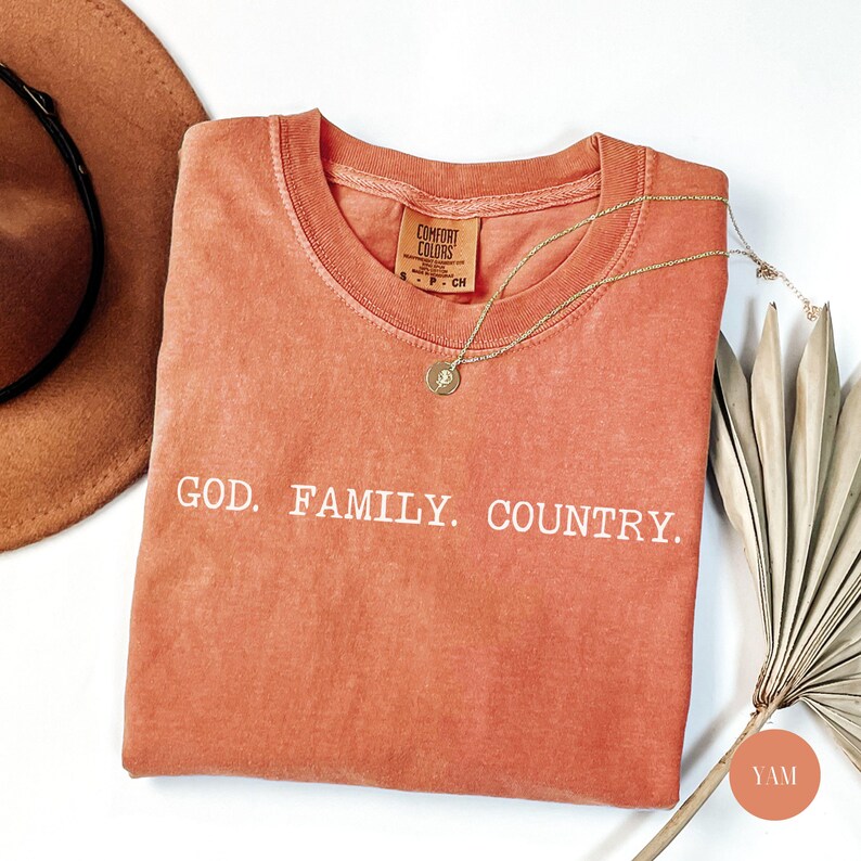 May include: A coral-colored Comfort Colors t-shirt with the words "GOD. FAMILY. COUNTRY." in white. A gold necklace with a pendant rests on the shirt. A brown hat and dried palm leaves are in the background.