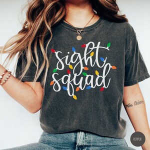 Puede incluir: Una camiseta gris oscuro con el texto "sight squad" en letras blancas. El texto está rodeado de luces navideñas de colores.
