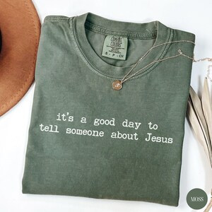 K&ouml;nnte beinhalten: Olivgr&uuml;nes T-Shirt mit dem wei&szlig;en Text "it's a good day to tell someone about Jesus". Eine goldene Halskette mit Anh&auml;nger liegt auf dem Shirt. Das Shirt hat einen Rundhalsausschnitt und ein Comfort Colors-Etikett.