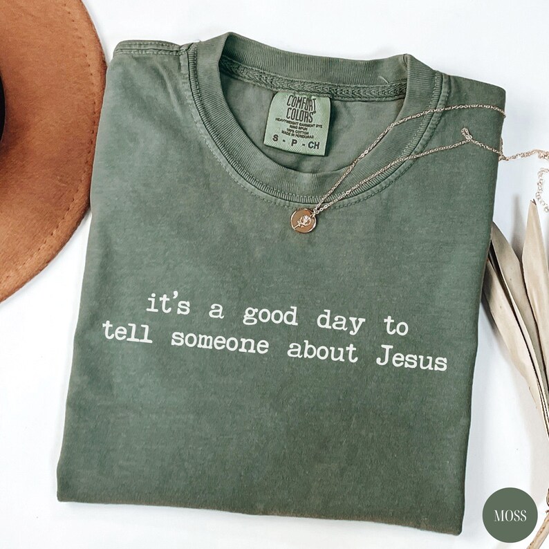 K&ouml;nnte beinhalten: Olivgr&uuml;nes T-Shirt mit dem wei&szlig;en Text "it's a good day to tell someone about Jesus". Eine goldene Halskette mit Anh&auml;nger liegt auf dem Shirt. Das Shirt hat einen Rundhalsausschnitt und ein Comfort Colors-Etikett.