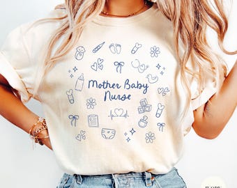 Op de afbeelding: Een wit T-shirt met een blauw grafisch ontwerp met de tekst "Mother Baby Nurse" omringd door babygerelateerde pictogrammen. Het shirt is verkrijgbaar in ivoor.