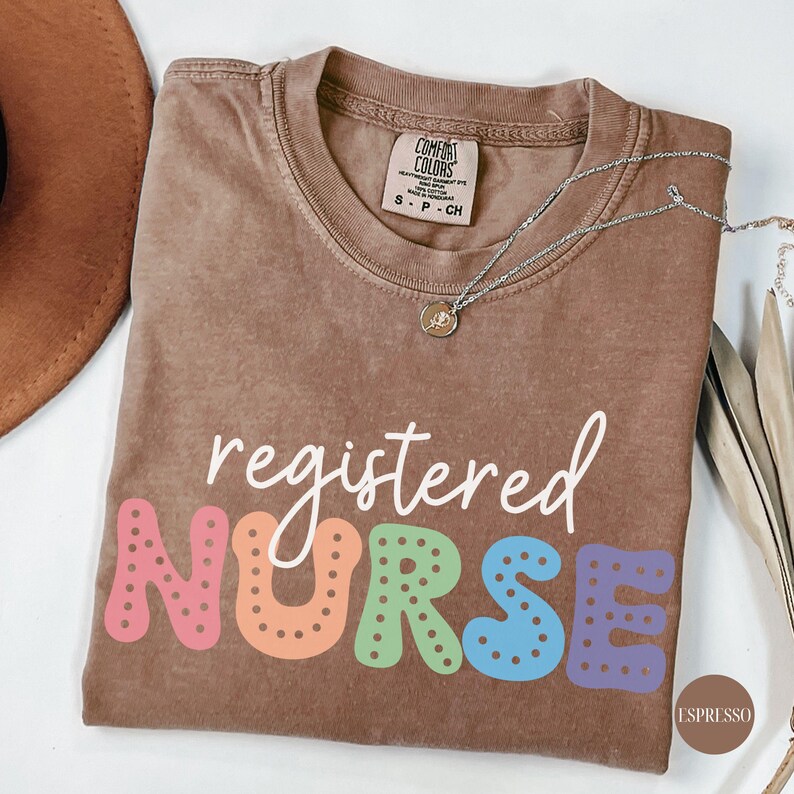 May include: A brown t-shirt with the text "registered NURSE" in colorful letters. The letters are outlined in white and have white polka dots. The shirt is made of a soft, comfortable material.