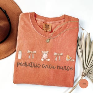 May include: A peach-colored t-shirt with the text "pediatric cricu nurse" printed on it. The shirt features a baby bottle, a teddy bear, and a pacifier.