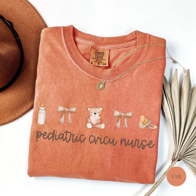 May include: A peach-colored t-shirt with the text "pediatric cricu nurse" printed on it. The shirt features a baby bottle, a teddy bear, and a pacifier.