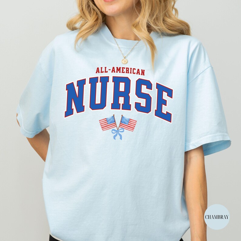 Op de afbeelding: Lichtblauw T-shirt met de tekst "ALL-AMERICAN NURSE" in rood en blauw. Twee Amerikaanse vlaggen zijn eronder gekruist. Het shirt heeft korte mouwen en een ronde hals.