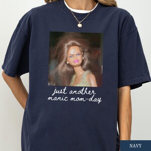 May include: Navy blue t-shirt featuring a framed image of a doll with a displeased expression. The image is accompanied by the text "just another manic mom-day" in a handwritten style font. The shirt has short sleeves and a crew neck.