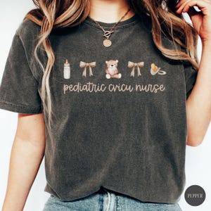 May include: A dark gray t-shirt with the text "pediatric cricu nurse" and images of a baby bottle, two bows, a teddy bear, and a pacifier.