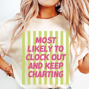 Op de afbeelding: Cr&egrave;mekleurige sweatshirt met de tekst "MOST LIKELY TO CLOCK OUT AND KEEP CHARTING" in roze letters, tegen een achtergrond van groene verticale strepen. De trui heeft een ronde hals.