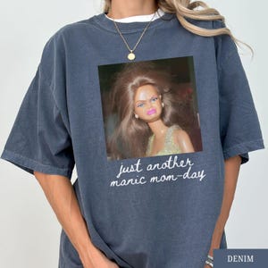 May include: Denim blue t-shirt featuring a framed image of a Barbie doll with a displeased expression. The text "just another manic mom-day" is printed below the image in a cursive font. The shirt has short sleeves and a crew neck.
