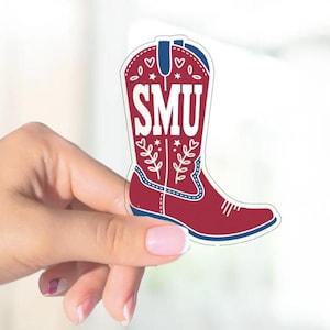 May include: A maroon and blue cowboy boot sticker with the letters "SMU" in white. The boot features decorative elements like hearts, stars, and floral designs. The sticker is held by a hand.