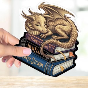 May include: A golden dragon with closed eyes is curled up asleep on top of a stack of four books. The books have titles that include "Fourth", "Iron Flame", "Onyx Storm", and "King".