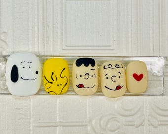 May include: Five fake nails with cartoon characters painted on them. The nails are arranged in a row on a white background. The characters are Snoopy, Woodstock, Charlie Brown, Linus, and a heart.