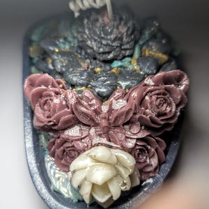 May include: A decorative candle in a dark grey oval container, featuring a central wick surrounded by dark grey textured wax, a central butterfly figure, two mauve rose-shaped wax accents, and a single white succulent-shaped wax piece at the base.