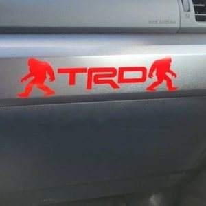 May include: A red TRD decal with Bigfoot silhouettes on a vehicle dashboard. The decal is centered on a gray panel, with the TRD letters in bold red. The dashboard is black, with various controls and vents visible.